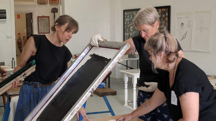 A woman looks on holding an ink squeegee while another lifts a silkscreen frame up and a third holds a piece of paper