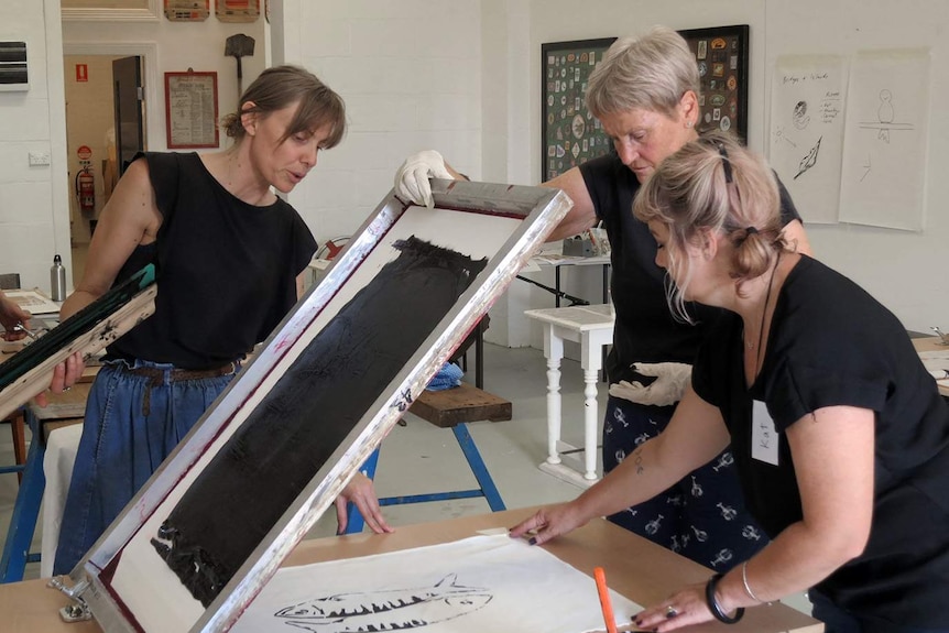 A woman looks on holding an ink squeegee while another lifts a silkscreen frame up and a third holds a piece of paper