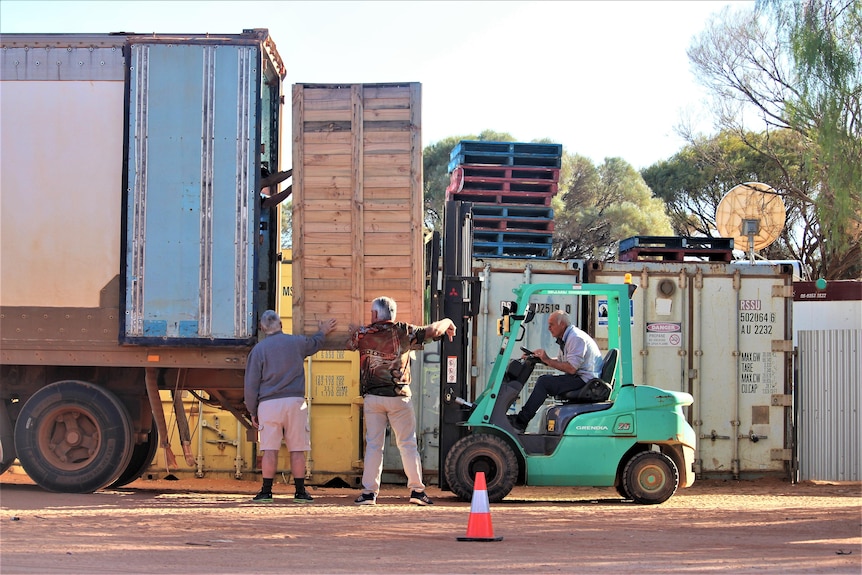 Two men help balance a crate on a forklift, another man drives the forklift 
