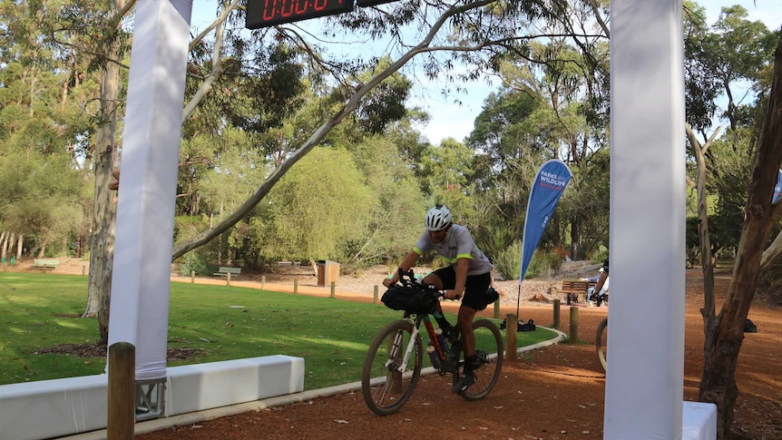 Declan von Dietze rides his mountain bike across a finish line in a park with a clock above reading 04:06:39:47.