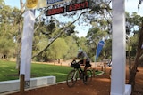 Declan Von Dietze rides his mountain bike across a finish line in a park with a clock above reading 04:06:39:47.