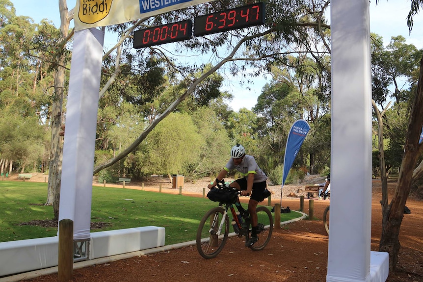 Declan von Dietze rides his mountain bike across a finish line in a park with a clock above reading 04:06:39:47.