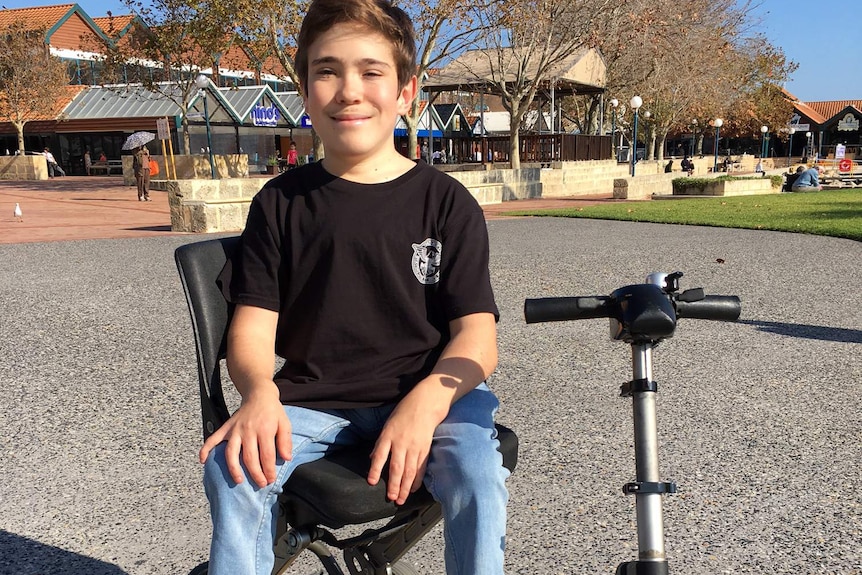 Coby Antonio sits in a yellow motorised scooter wearing a black t-shirt, jeans and black shoes.