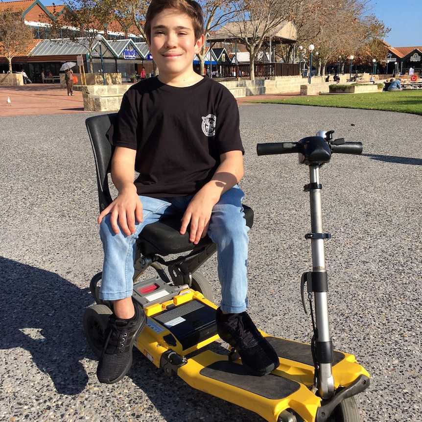 Coby Antonio sits in a yellow motorised scooter wearing a black t-shirt, jeans and black shoes.