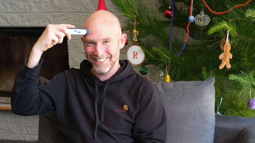 A man smiles at the camera while holding a device to his forehead to test his temperature