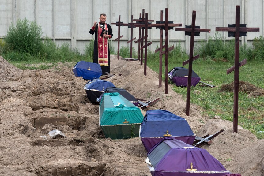 A priest stands behind a line of covered coffins placed beside open graves