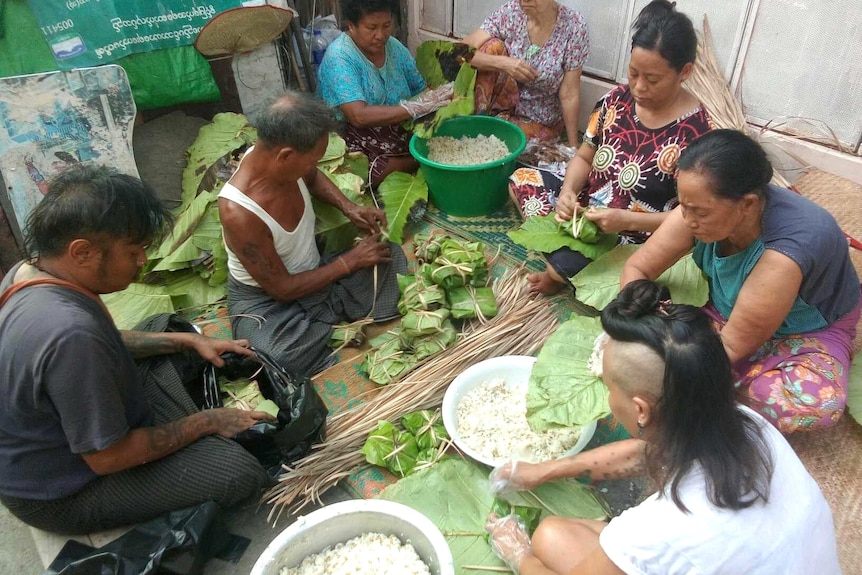 A group of men and women sit on the ground and wrap rice in a green leaf.