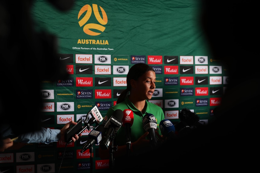 Microphones surround a woman who's speaking in front of a branded backdrop