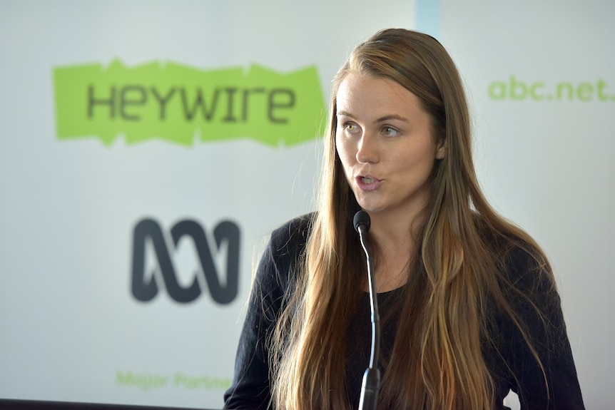 A woman with long hair is standing at a lectern with the words Heywire on a poster in the background