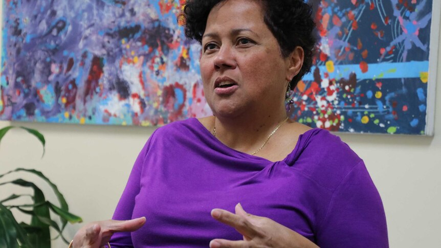 A woman in a purple dress speaks in front of a colourful painting.