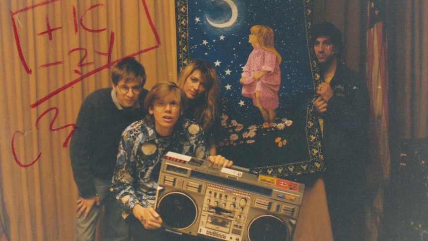 Sonic Youth stand in a wood panelled room. Thurston Moore holds a boombox, Lee Ranaldo holds a hanging rug.