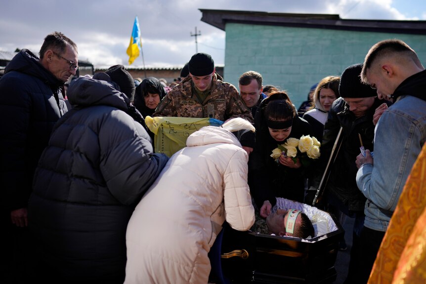 A group of people, holding flowers and Ukrainian flags, cry over the body of a soldeir lying in a coffin.