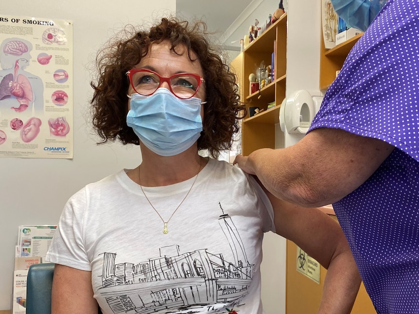 A masked, bespectacled woman with shoulder-length, curly hair getting a COVID jab.