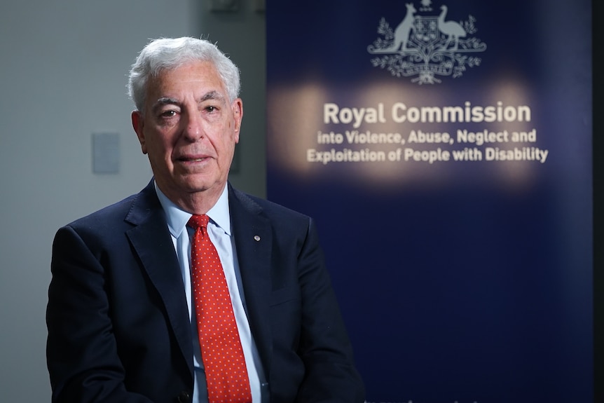 An older white man with a suit and tie sitting in front of a disability royal commission sign