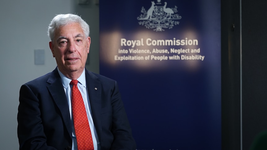 An older white man with a suit and tie sitting in front of a disability royal commission sign