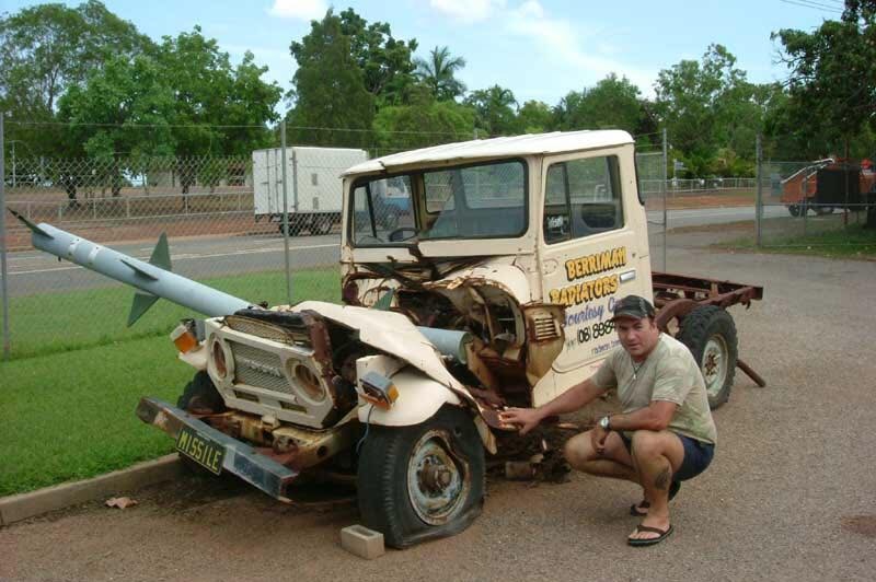 A man crouches beside a ute with a bumper crumpled under a missile.