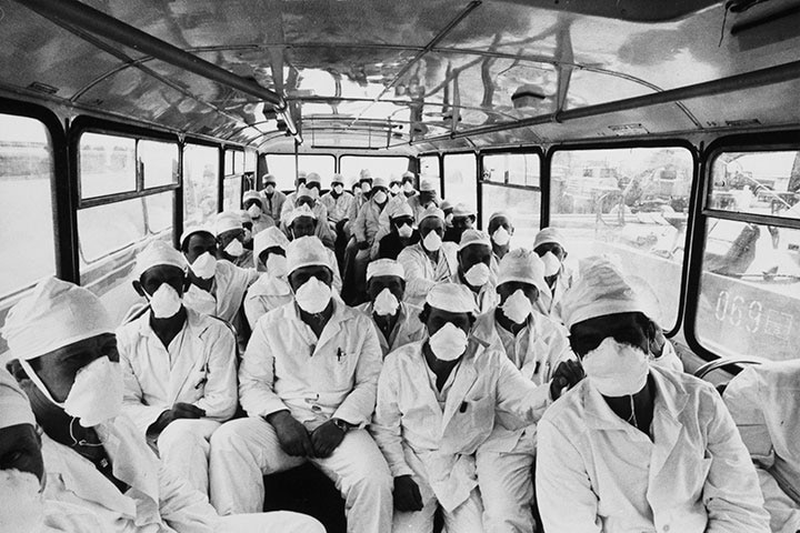 A group of scientists in white coats, hats and masks sit crowded in a bus