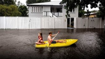 Two children paddle a kayak on a flooded street  in front of a Townsville home.
