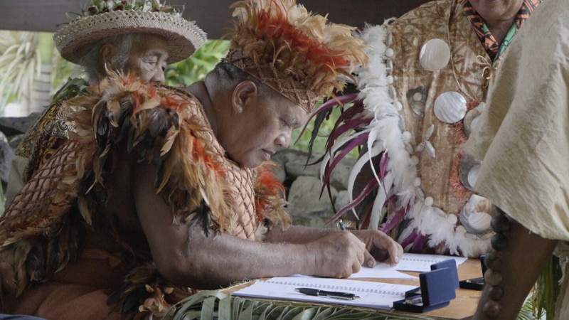 Man in traditional dress signs treaty to protect whales. Elders in traditional dress surround him. 