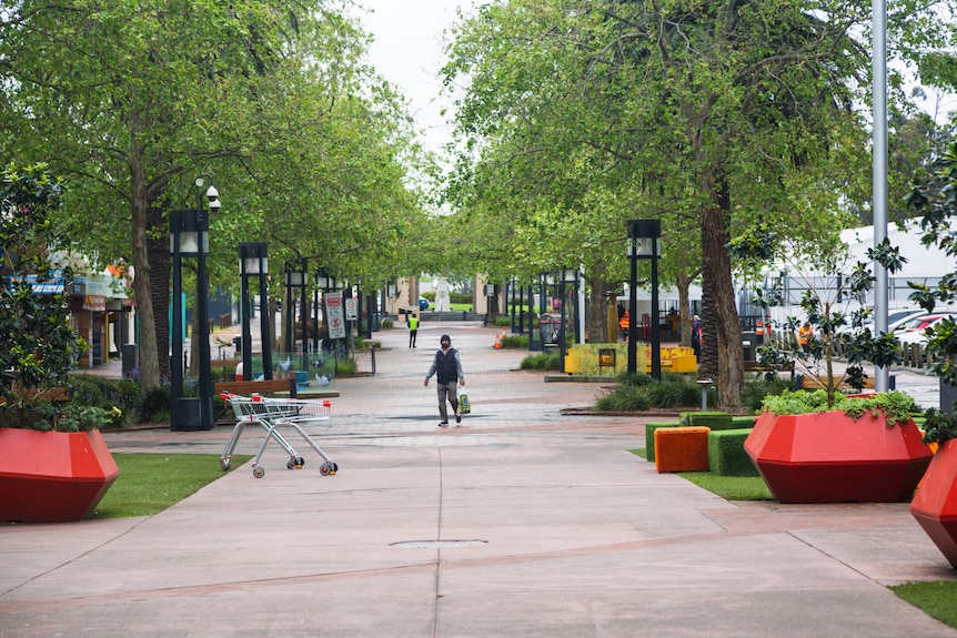 A person wearing a face mask is a small figure in the distance of an empty plaza with leafy trees during lockdown.