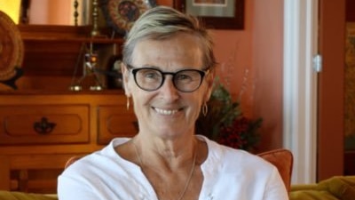 Woman with glasses sits on a couch looking directly at camera and smiling.