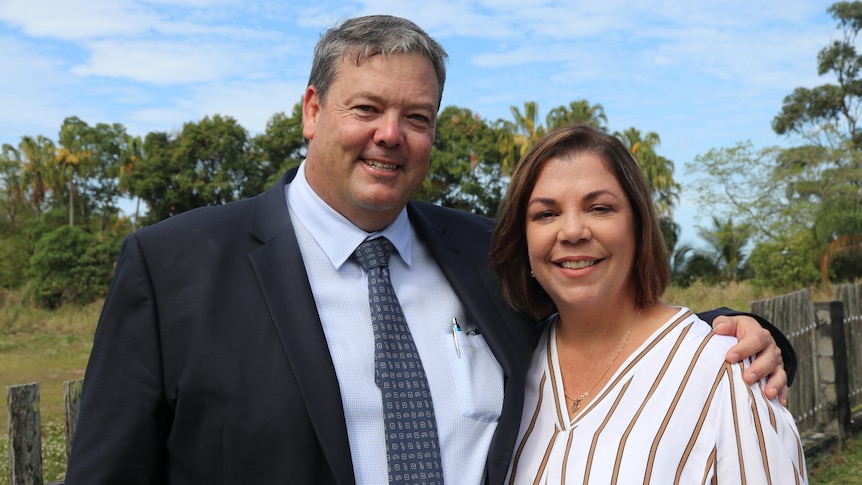 A middle aged man stands in front of a fence with his arms around his wife