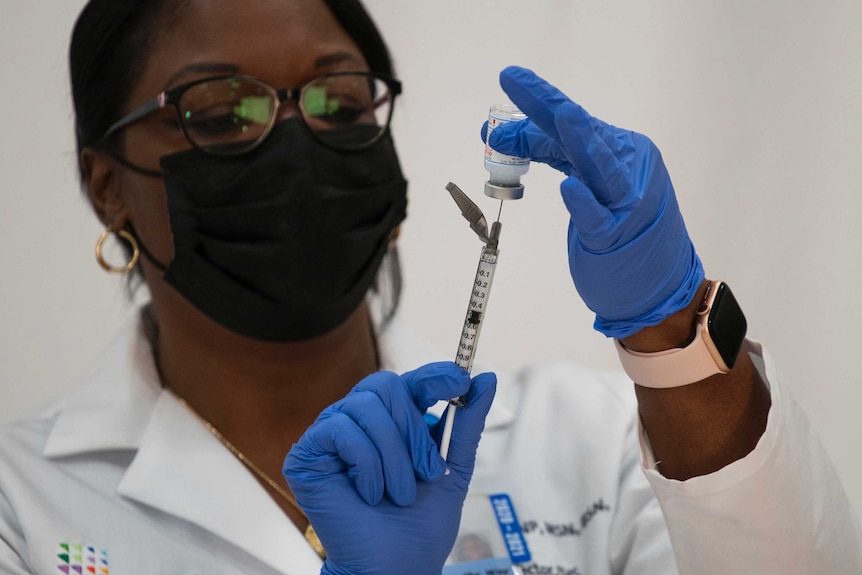 A woman wearing a blue mask prepares a dose of vaccine from a vial.