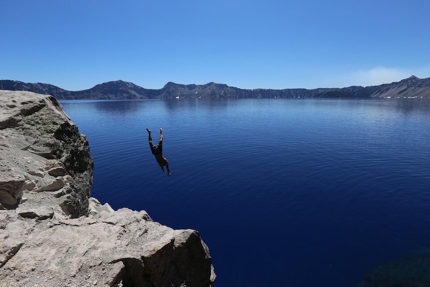 A man diving off a rock cliff into a blue lake with mountains in the background 