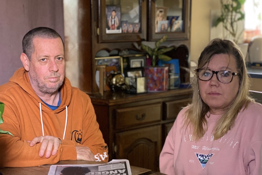 a man in an orange hoodie and a woman in a pink sweatshirt sitting a table inside a home looking grim