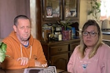a man in an orange hoodie and a woman in a pink sweatshirt sitting a table inside a home looking grim