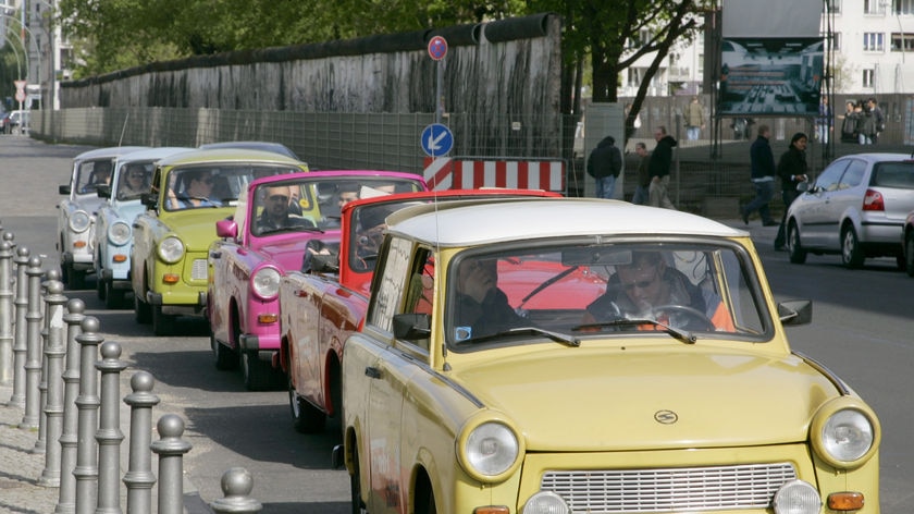 Trabant cars drive in front of the remains of the 'Mauer' frontier wall