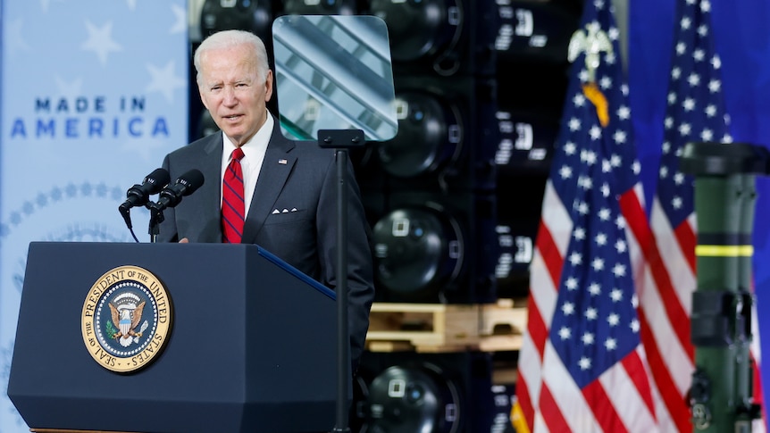 Joe Biden stands at a lectern at a weapons factory in front of a sign that says made in America. 