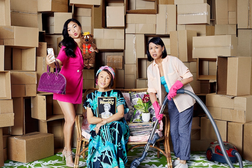 Three women pose on set with cardboard box wall, one takes selfie, another sits smiling with magazine as one holds vacuum.