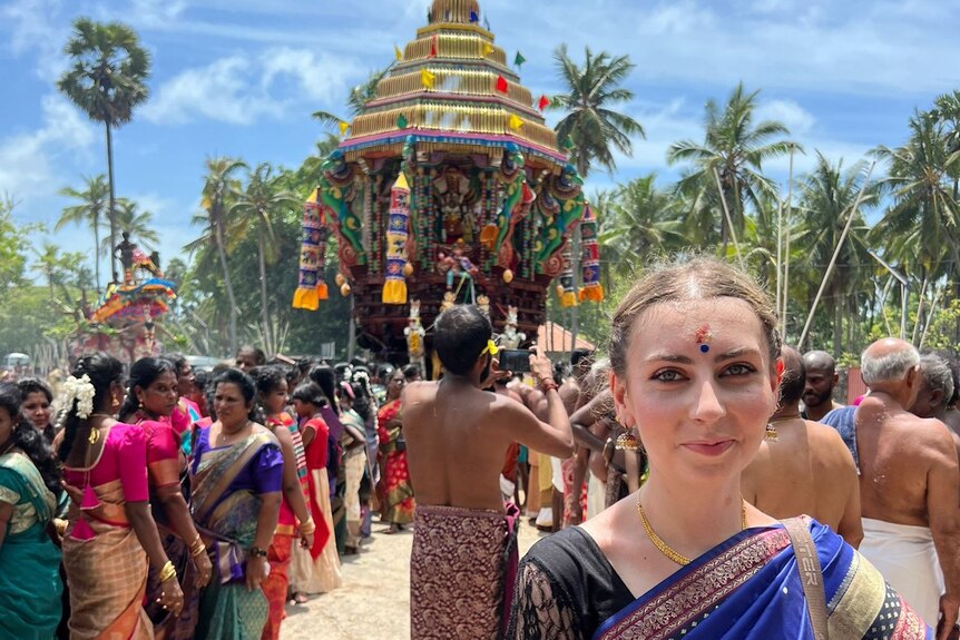 A woman stands in front of a temple and palm tree and colourful crowds.
