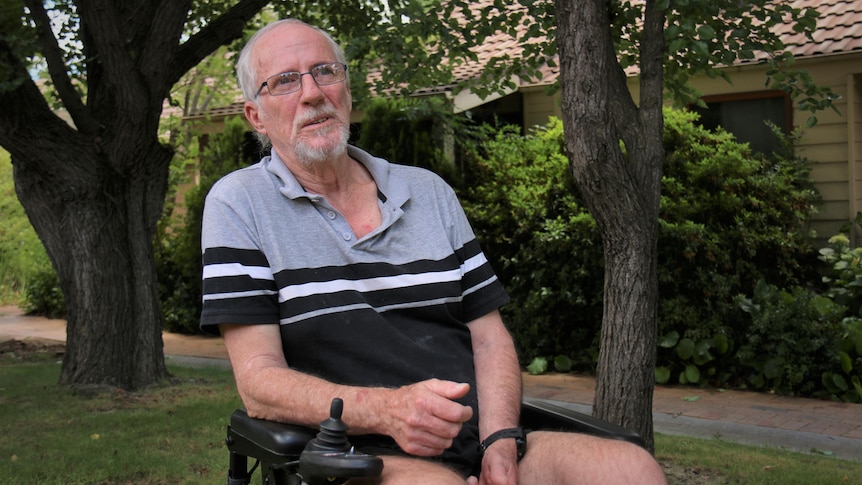 A man sits in an electric wheelchair on the front lawn of a house with trees behind him