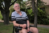 A man sits in an electric wheelchair on the front lawn of a house with trees behind him