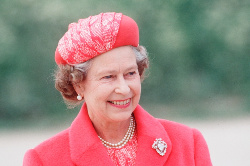Queen Elizabeth smiles, wearing a pink hat and matching coat with an ornate diamond brooch on the lapel