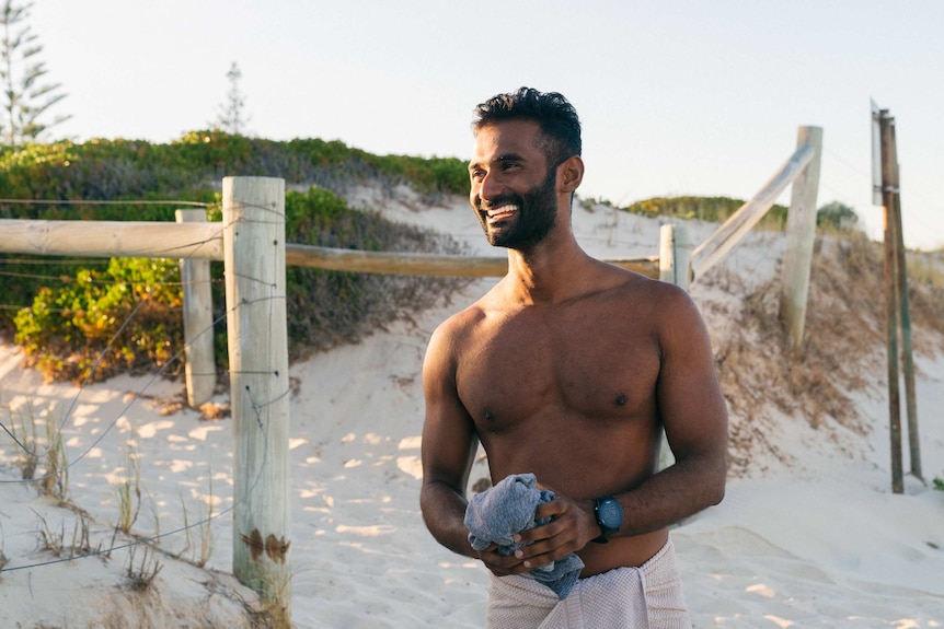 A man wearing only a towel smiles while standing at a beach