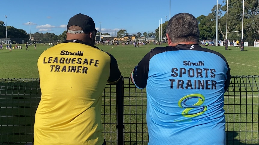 Two men watch a game of football wearing shirts that read "TRAINER"