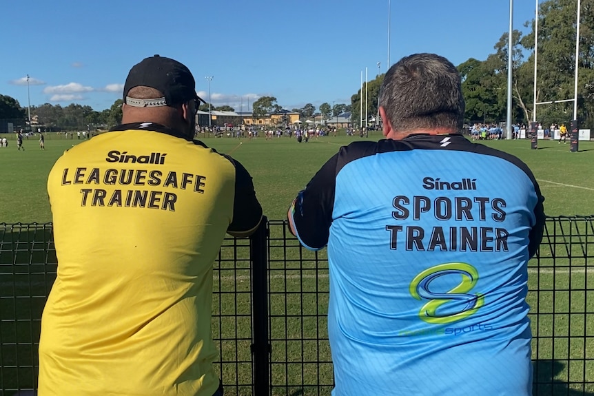 Two men watch a game of football wearing shirts that read "TRAINER"