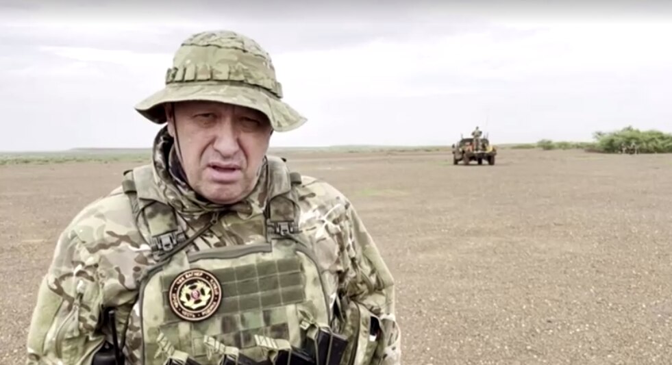 A man in camouflage gear stands in a deserted landscape with a truck far in the background