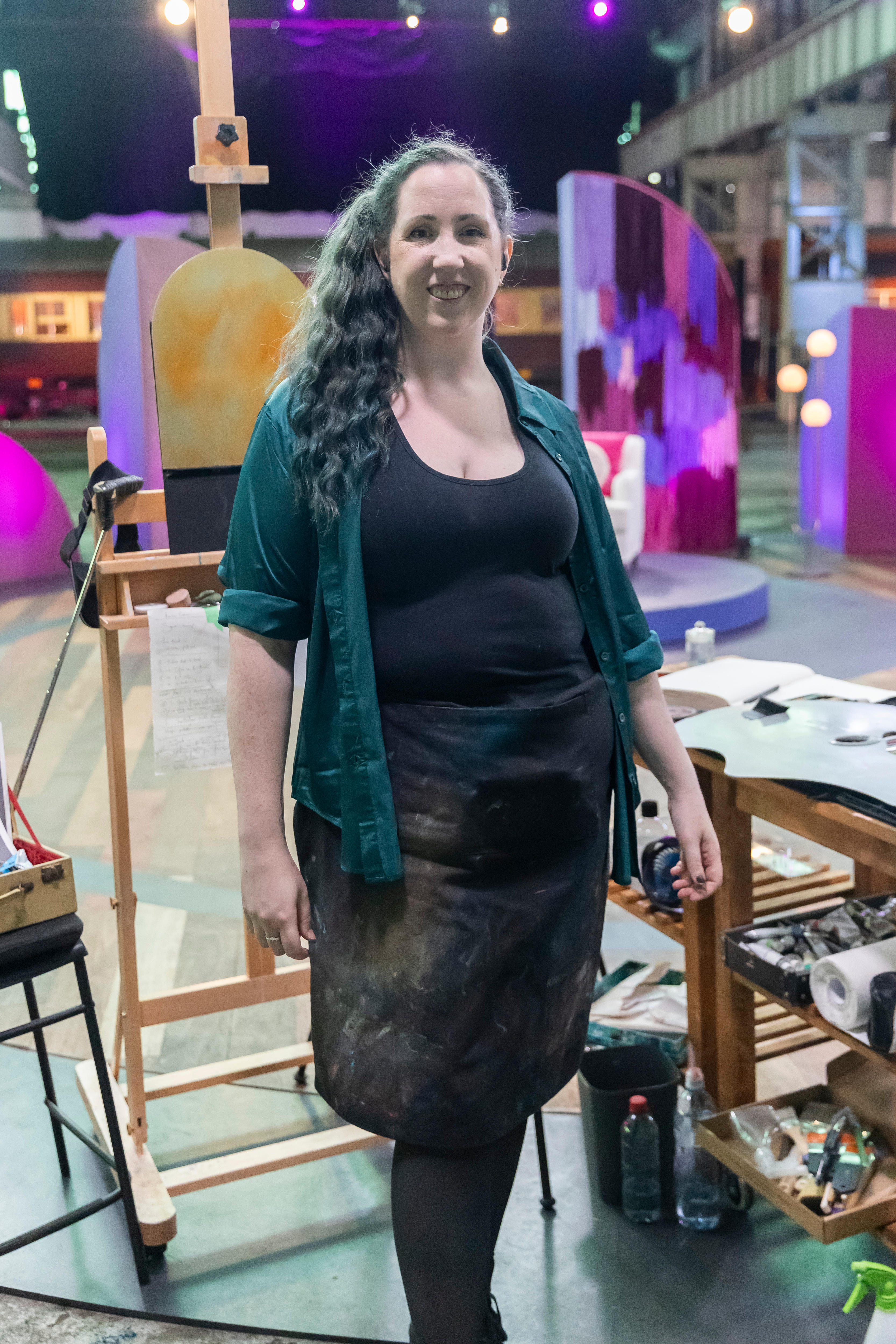 A woman with long brown curly hair smiles at the camera. Behind her is an easel