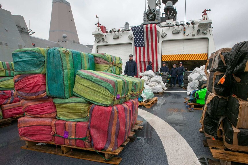 large packages wrapped in red, green and black plastic are loaded on pallets on a ship with crew and a US flag on it