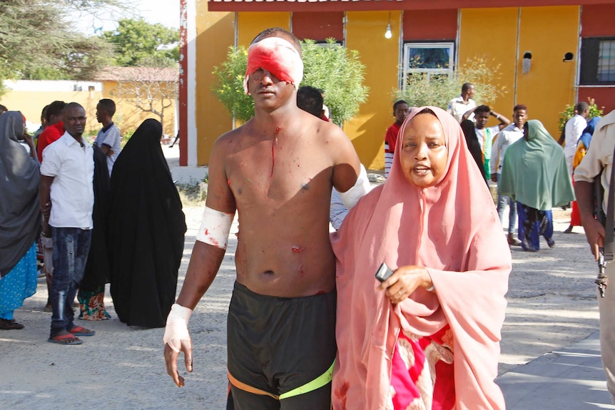 A woman in a hijab leads a man with no shirt who has a bloodied bandage on his head.
