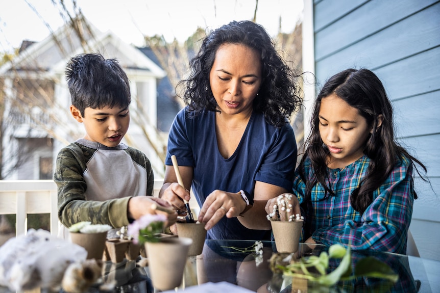 A woman and two children either side of her work with small pots and plants on a table outside.