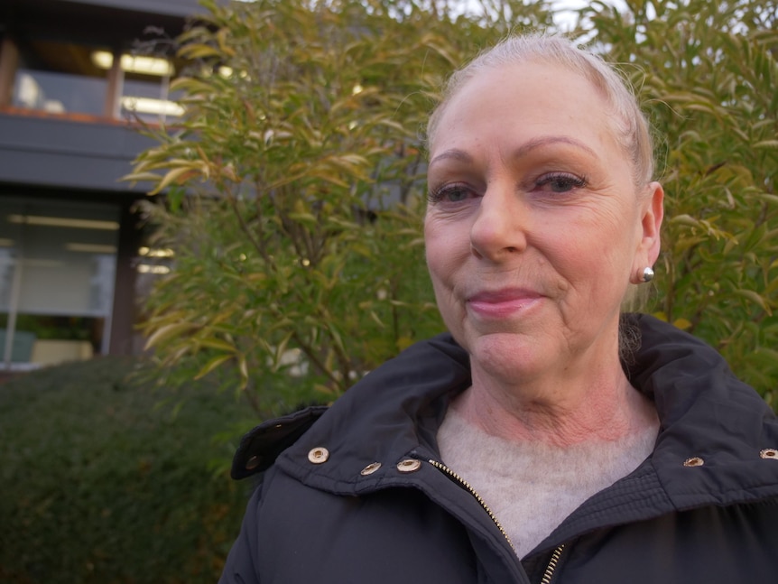 A woman with tied back hair smiling to camera, with greenery and a building behind her.