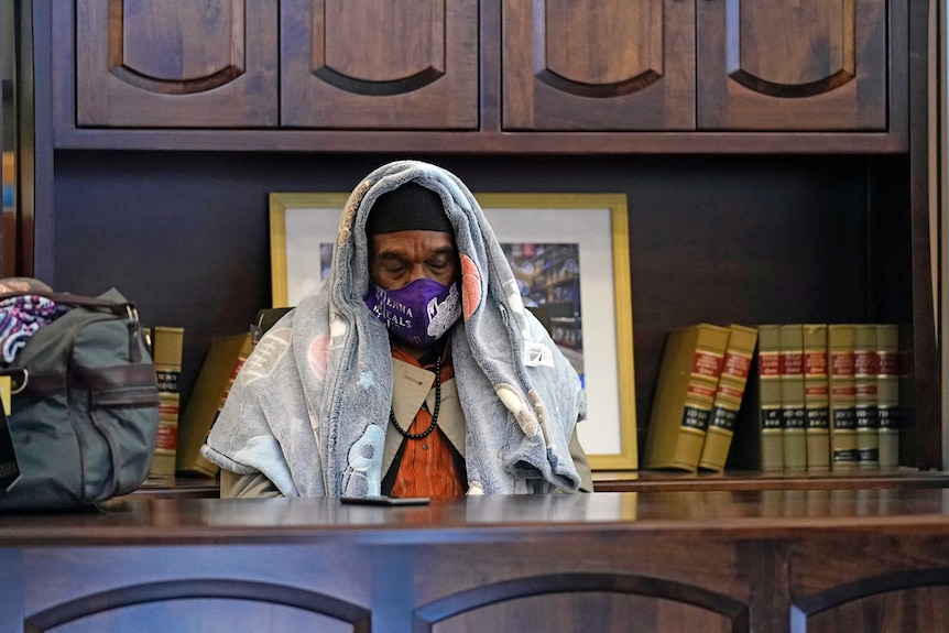 A man in a face mask with a blanket on his head sits behind a desk