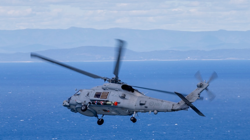 A military helicopter tilts towards land as it flies over ocean.