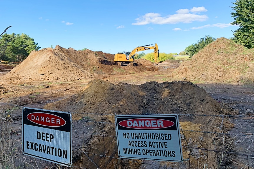 An excavator digs behind a fence with warning signs