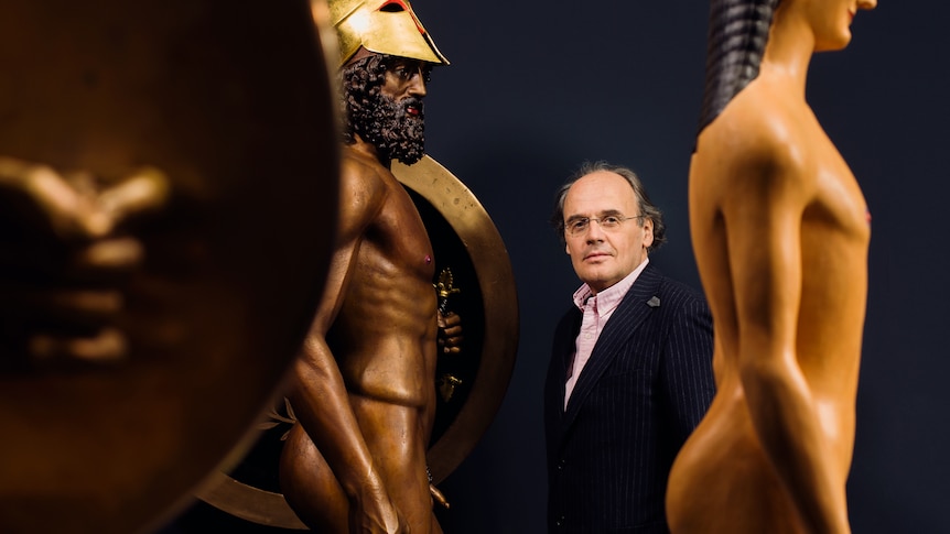 A white man in his 60s wearing glasses and suit stands between two golden statues